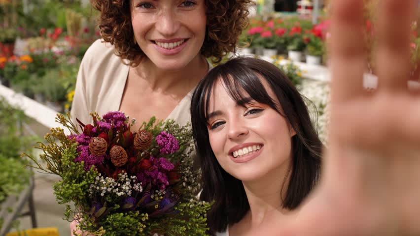 Two florists are posing, holding a colorful bouquet of flowers and smiling at the camera in their greenhouse