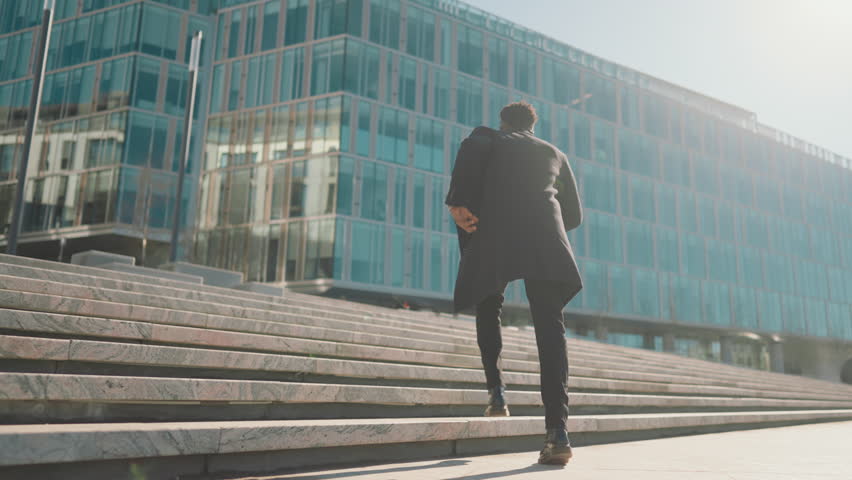 Young black man going up on stone staircase in city, back view, move to goal. Slow motion shot outdoors in downtown, businessman running to office center, be late and motion to dream, career growth