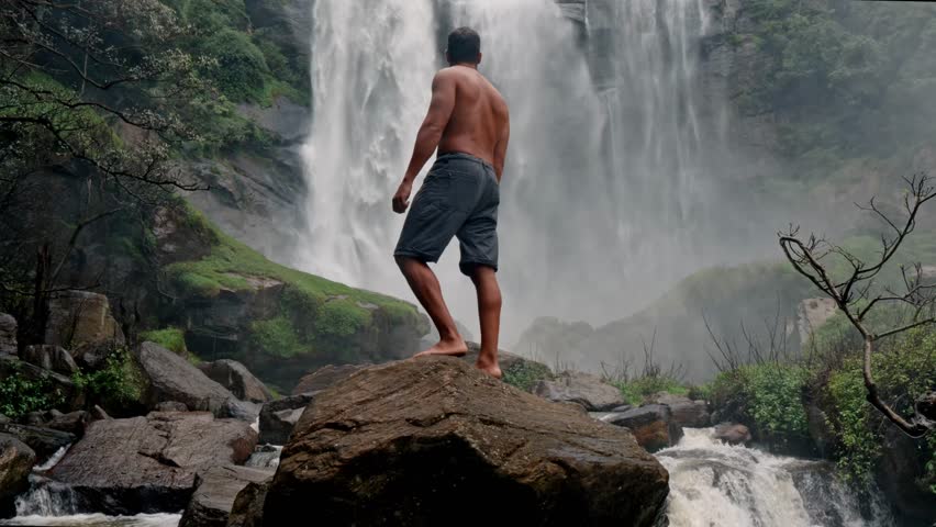 A man stands near the base of Bomburu Ella Waterfalls, surrounded by mist, rocks, and dense greenery in the highlands of Nuwara Eliya, Sri Lanka.