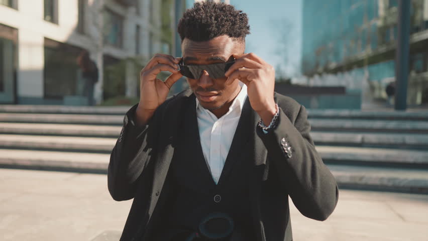 Cool black guy putting fashionable sunglasses on face, portrait of stylish man. Handsome male passerby sitting in park area in downtown, trendy style on city streets, african american male person