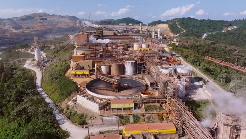 Barrick Gold mine complex showing large scale concentrators, tanks, pipes, steam vents, and surrounding hills in Cotuí Republic. Aerial flyover