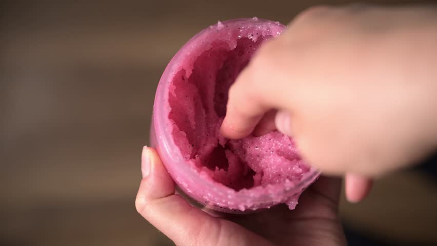 Female hand scoops pink raspberry sugar body scrub from jar showing its texture. Smooth, creamy texture ideal for exfoliating and nourishing skin. Body scrub for skincare, wellness and spa concepts.