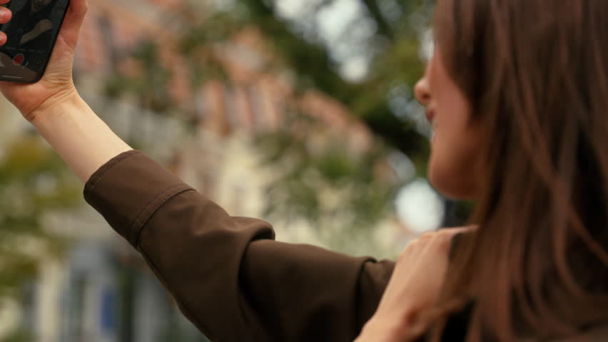 Back view from behind Caucasian woman female businesswoman business girl in city holding mobile phone smartphone distant call record video vlog blog recording take pictures selfie photo influencer app