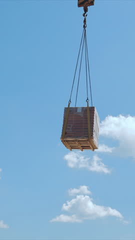 Crane lifting pallet of bricks at construction site, A crane lifting a pallet of bricks high into the air at a construction site, set against a clear blue sky with some clouds