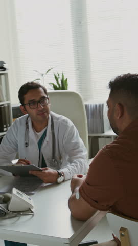 Vertical shot of clinician with medical card of patient looking through information and consulting man talking about his health and pointing at glucose monitoring system patch on his arm