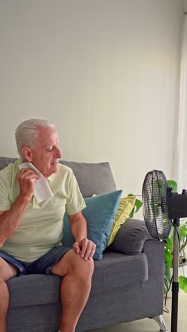 Vertical video of Senior man refreshing himself with an electric fan and wiping sweat with a tissue