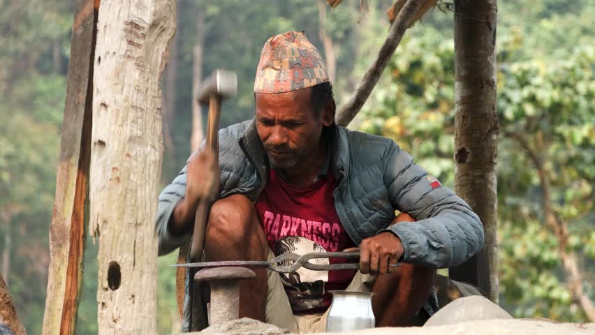 An old man is making a sickle in a traditional village blacksmith workplace. He heats the metal and shapes it with a hammer. Great for videos about rural life, tradition, or handmade tools.