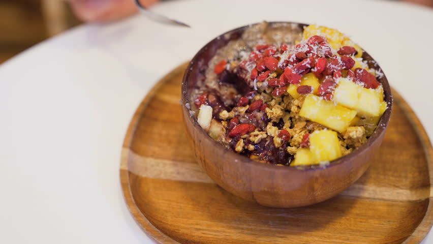 Close-up of wooden bowl with delicious acai bowl, topped with pineapple, mango, goji berries, granola and coconut flakes, being enjoyed by a customer.