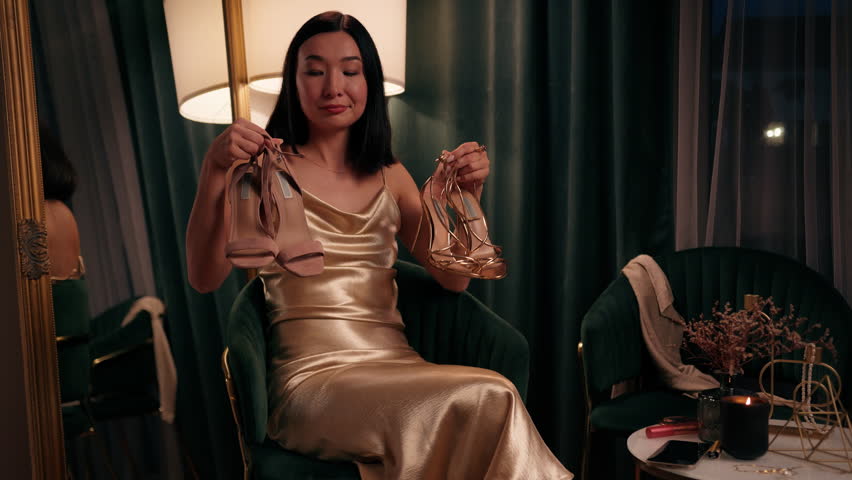 Beautiful stylish pensive unsure hesitate Asian Korean gorgeous woman Chinese lady Japanese girl female in golden dress style at home evening choosing between two pairs of shoes high heels choice