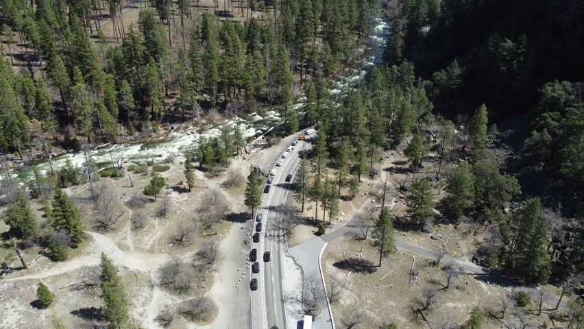 A Fast Moving River Stream Winding Through The Valley Floor Of Yosemite National Park. Spring To Summer Tourist Season, Slow Aerial Tilt Up Towards The Famous Granite Mountain Skyline.