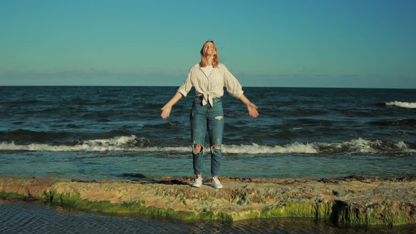 Blonde woman stretching arms on moss-covered seaside rock, wearing white shirt and ripped jeans, embodying natural freedom with expansive gesture