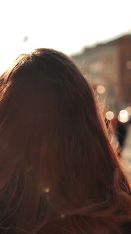 Sexy girl with long red hair turning around and smiling on camera outdoor, blurred city crowd at sunset on background with super slow motion. Portrait close-up view. Vertical shot.