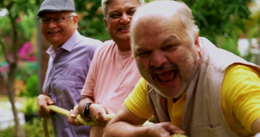 Senior playing tug of war in garden, Indian Asian elderly friends enjoying outdoor physical activity with rope, laughing and pulling together as part of fun reunion game amid green nature