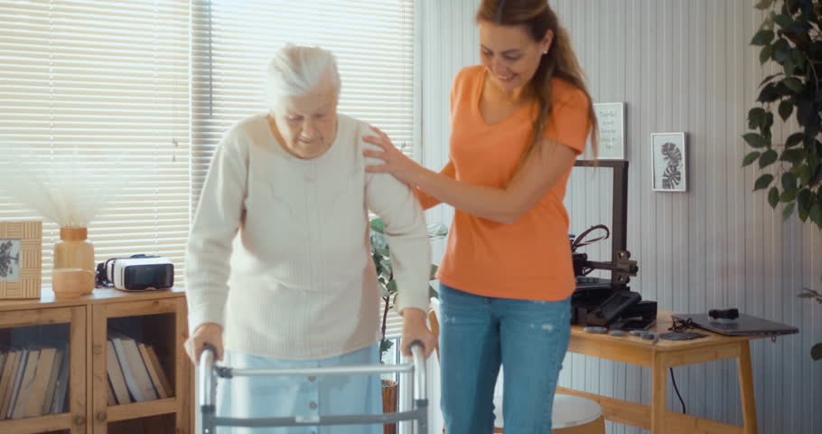 Woman volunteer, or adult daughter, helps an old 90s grandmother walk using a walker in the living room at home.