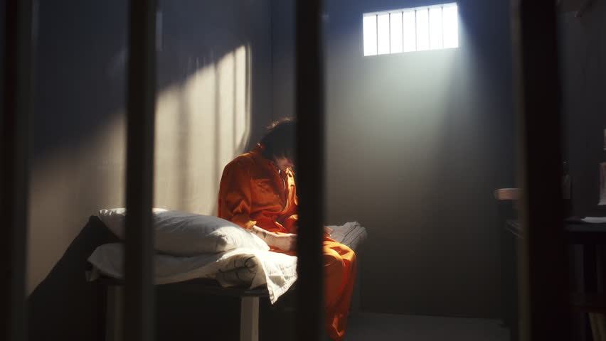 Young male inmate in orange uniform sitting on bed in prison cell, looking at barred window pensively and writing notes on paper
