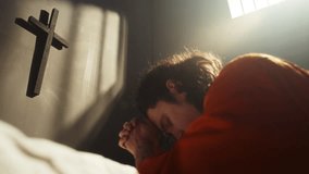 Inmate in orange uniform clasping hands and praying, looking at cross mounted on wall inside prison cell with warm sunlight streaming through barred window - Powered by Shutterstock - Get 15% off with code: PIKWIZARD15
