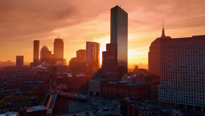 Skyscrapers of Boston skyline colored orange at sunset. Drone footage over the dark city at dusk time.