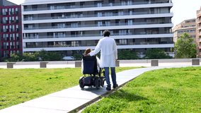 Doctor assisting a female patient in a wheelchair, strolling through a park adjacent to a modern hospital building - Powered by Shutterstock - Get 15% off with code: PIKWIZARD15