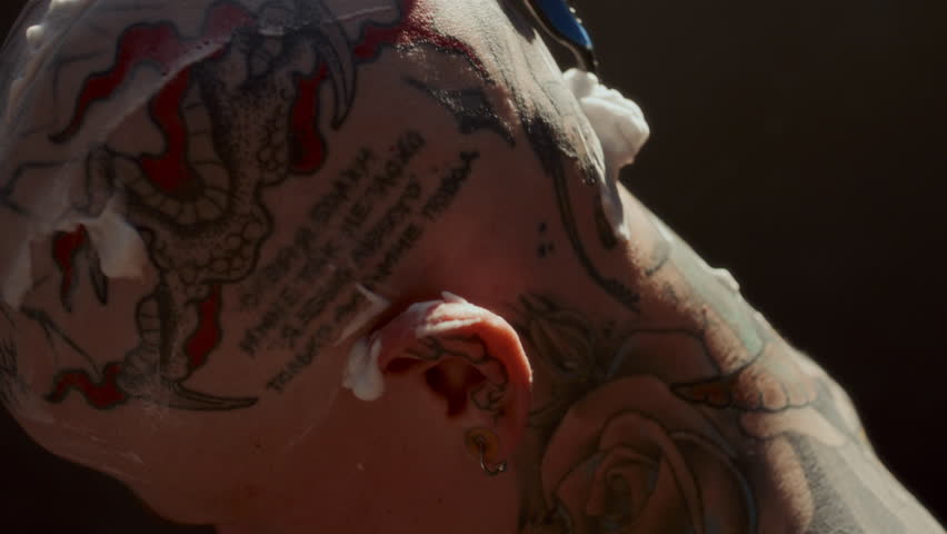 Man using razor as shaving his tattooed head covered with thick foam under warm sunlight. Close-up view