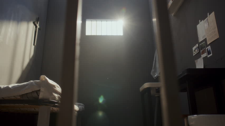 Shirtless bald man with tattoos exercising in confined prison cell by doing pushups on floor between bed and toilet. Zoom out shot through bars