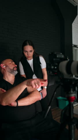 Bearded man in black clothes sits on the chair pointing at his camera. Brunette woman stands behind looking at equipment and nodding. Studio backstage. Vertical video.