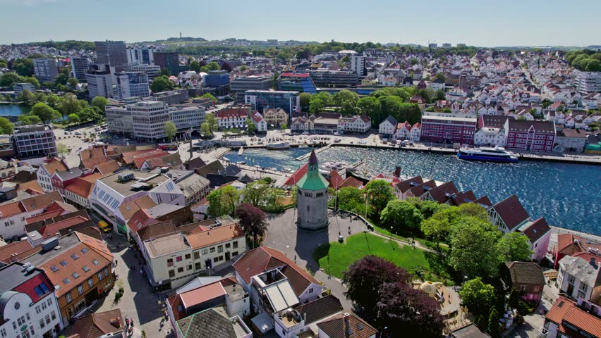 Drone circles Valberg Tower, showcasing its historic architecture with Vågen harbor and Stavanger cityscape in the sunny background.