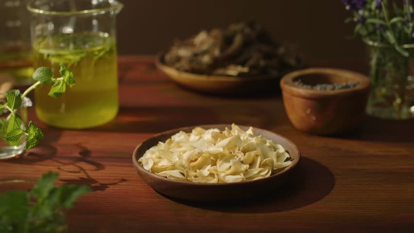 A hand from outside the frame reaches in and grabs a handful of white dried petals, raises in the air, and drops them back onto the plate. The petals fall, surrounded by bottles of essential oils.