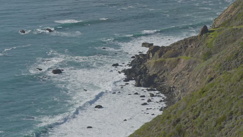 Aerial view of turquoise waves rolling against a steep, rocky shoreline along California’s iconic Big Sur coastline under soft midday light.