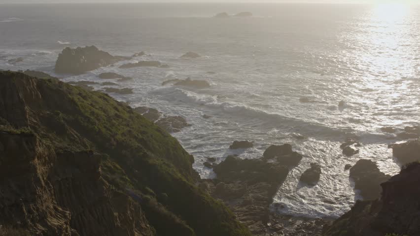 Dramatic golden hour light shines over Big Sur’s rugged coastal cliffs as ocean waves crash into jagged rocks below in this sweeping cinematic aerial shot.