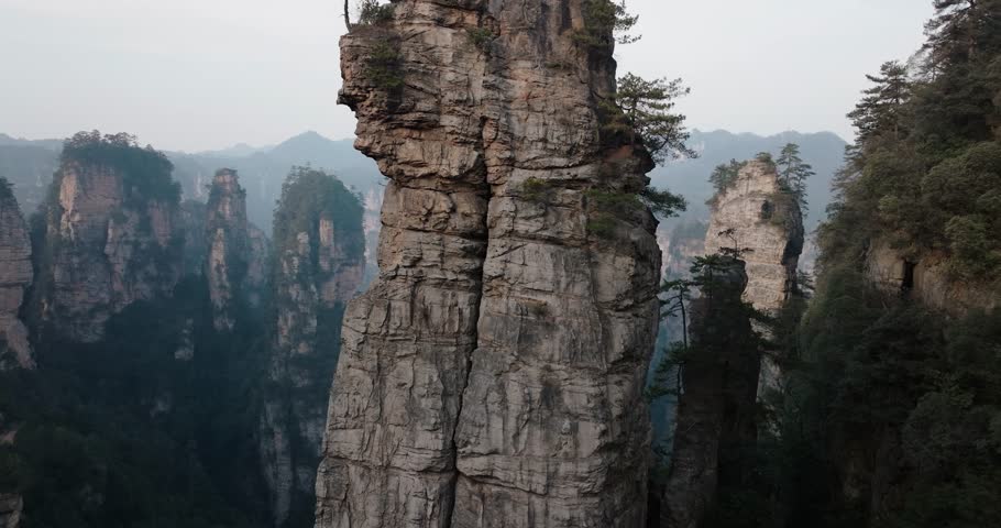 Aerial shot of a tall isolated sandstone tower with sparse trees on top, set against the dramatic forested cliffs of Zhangjiajie National Park, China