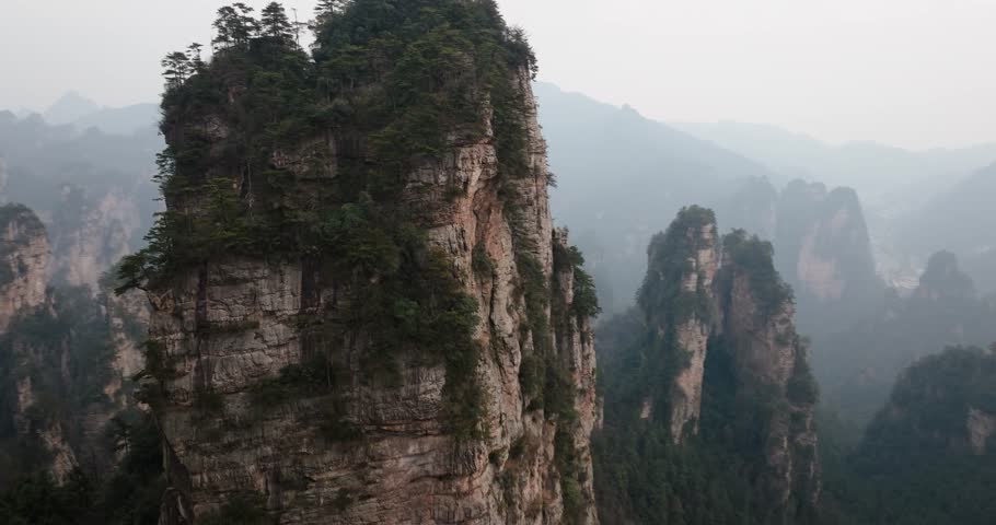 Orbiting aerial of a towering sandstone peak blanketed in trees with distant misty cliffs in Zhangjiajie National Park