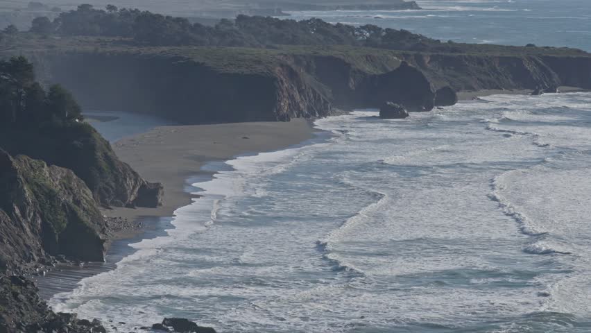 A wide, sweeping drone shot captures foamy waves rhythmically crashing onto the rugged shores of California’s dramatic coastline near Big Sur.