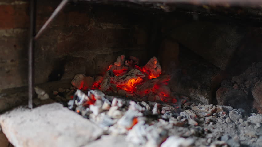 Close-up shot of glowing red-hot coals inside a traditional Argentine grill, with ash and embers prepared for asado barbecue, brick firebox visible, smoke rising gently, sliding and revealing.