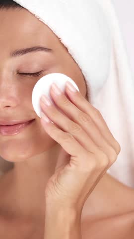 Young woman with radiant skin smiles while wiping her face with cotton pad. Ideal for self-care, skincare tutorials, clean beauty, minimalist wellness, and facial cleansing concepts.