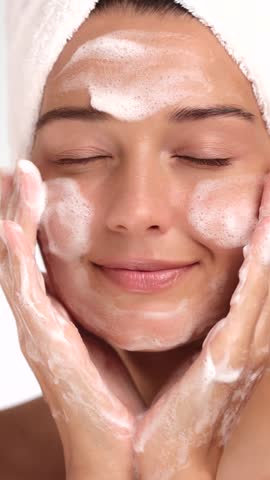 Caucasian woman in white towel gently cleanses her face with foamy cleanser. Ideal for skincare, beauty treatments, cosmetic ads, wellness content, and facial care promotional visuals.