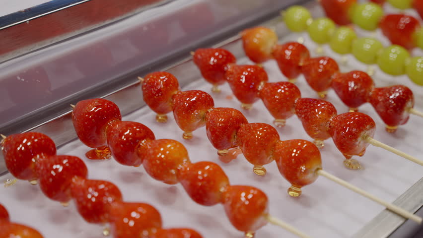 Rows of glossy, vividly colored candied fruit skewers displayed in close-up, showcasing glistening sugary coatings on strawberries, grapes, and other bite-sized fruits.