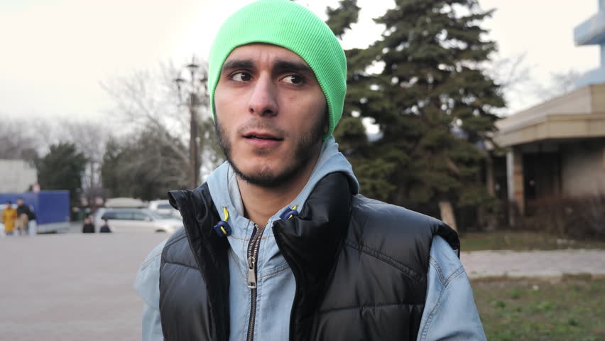 Caucasian man in sportive outfit engages in conversation standing on street in city. Relaxed bearded guy in green hat communicates with friends on weekend