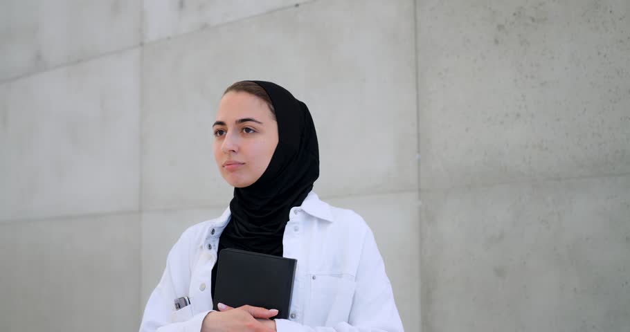 Young muslim woman in hijab leaning against concrete wall, holding book and gazing thoughtfully, reflecting on academic goals and professional aspirations