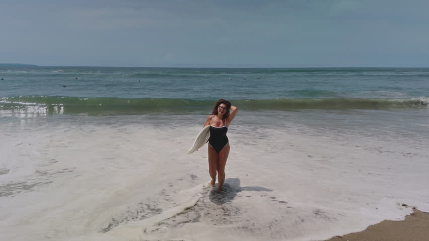 Active mature woman in wetsuit with surfboard at the beach, enjoying a healthy lifestyle. Old senior female doing surfing on Bali. Elderly lady holding surf board. Tropical coast in asia, big wave.