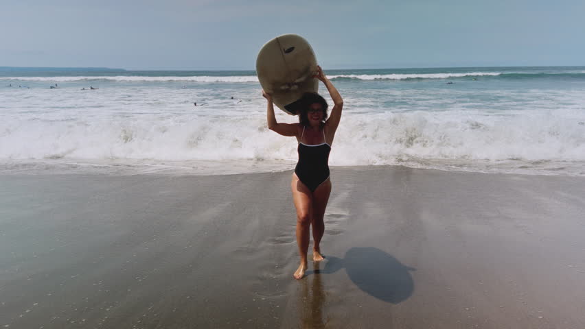 Active mature woman in wetsuit with surfboard at the beach, enjoying a healthy lifestyle. Old senior female doing surfing on Bali. Elderly lady holding surf board. Tropical coast in asia, big wave.