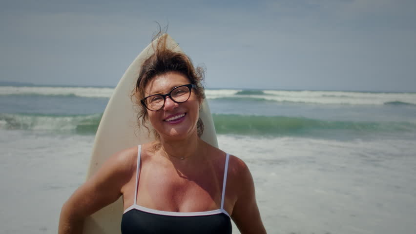 Active mature woman in wetsuit with surfboard at the beach, enjoying a healthy lifestyle. Old senior female doing surfing on Bali. Elderly lady holding surf board. Tropical coast in asia, big wave.
