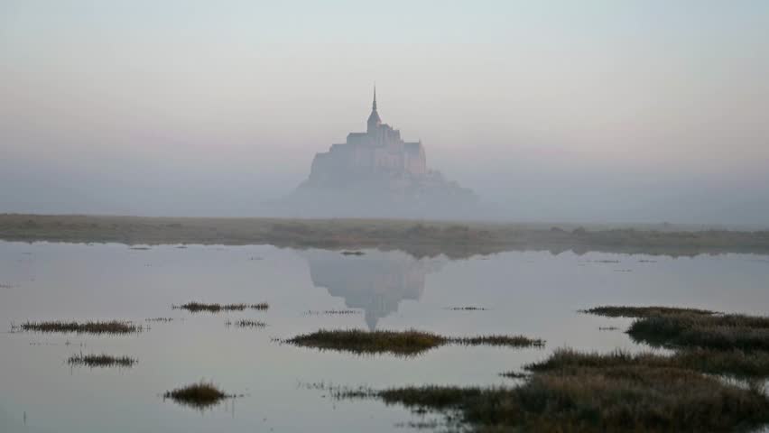 The iconic Mont Saint-Michel emerges through thick fog on a gloomy day, with its medieval village and abbey shrouded in mist on the Normandy coast