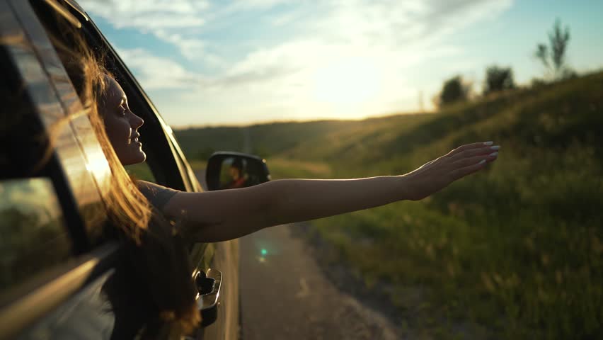 woman enjoys summer drive. Hand stretches out car window feeling warm breeze sunlight. Hair flows wind during golden hour. Carefree summer road trip embracing nature and freedom. Woman car hand summer