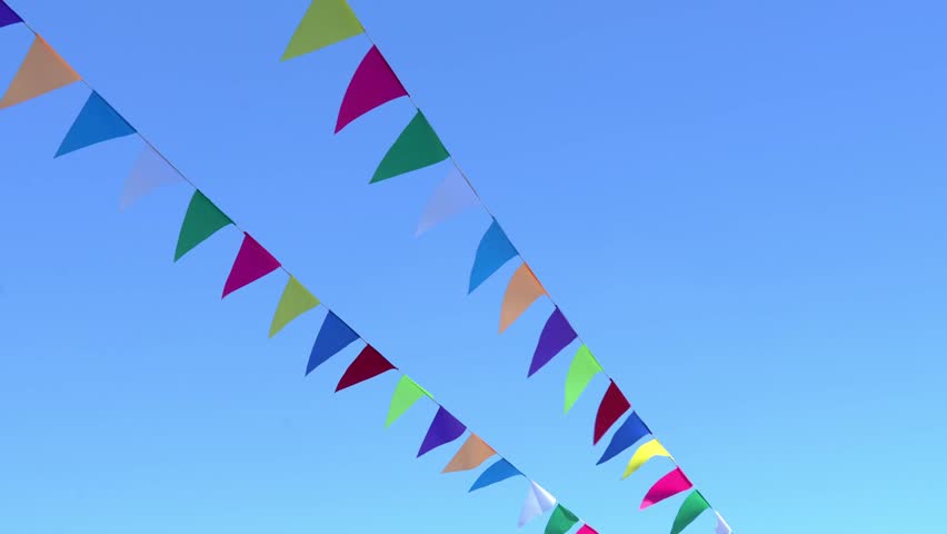 The wind spreads colorful flags against a clear sky. The image shows colorful triangular flags hanging against a clear blue sky. The flags form two rows, creating a feeling of festivity.