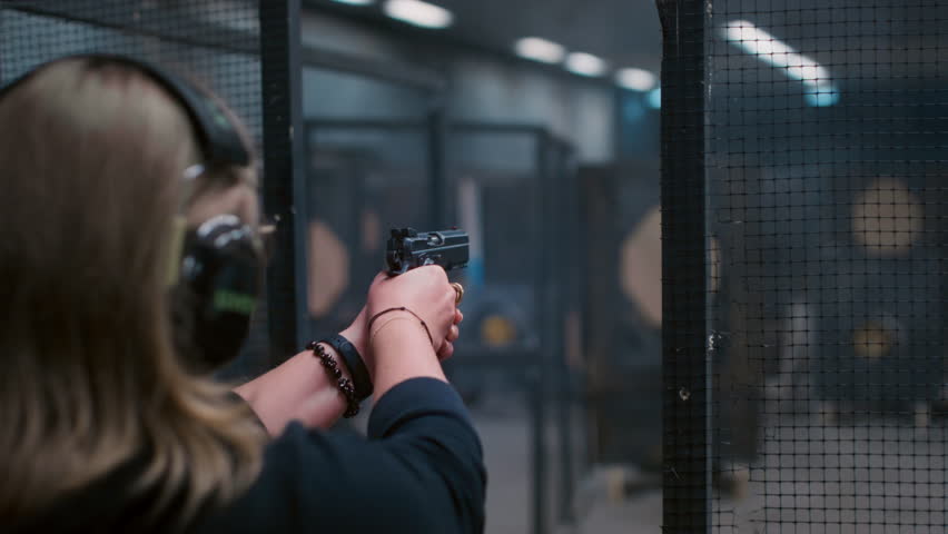 Woman with gun shoots a firearm at a shooting gallery or range. Female is engaged in sports shooting, wearing headphones. Pistol is a small weapon. Cartridge case goes off in slow motion