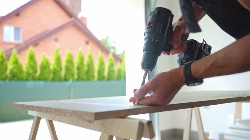 Man in work overalls bib using cordless power drill to make hole in laminated wood board. Male carpenter working in home workshop, drilling chipboard on workbench