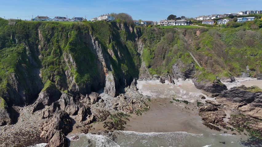 A drone view of houses on top of moss-covered cliffs on Polstreath Beach with calm waves on a sunny day in Mevagissey, Cornwall, United Kingdom