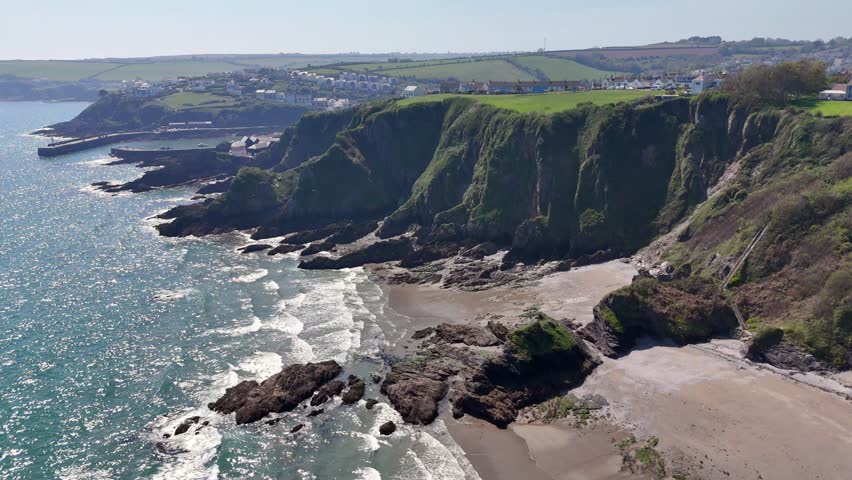 An aerial scenic view of Polstreath Beach, the waves crashing the green rocky cliffs, houses in the background on a sunny day in Mevagissey, Cornwall