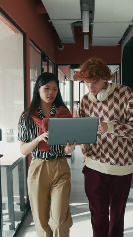 Vertical tracking shot of two diverse social media managers walking towards camera with laptop while discussing creation of new content, coworking space corridor