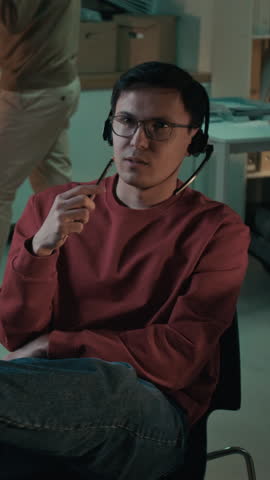 Medium vertical shot of Asian male scam caller relaxing on chair in office, rehearsing dialogue script lines, inventing creative social engineering approach to swindle victim